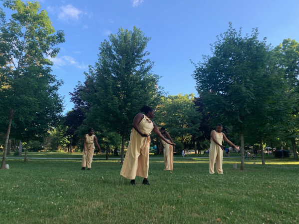 Four black women in matching beige shirts and pants dance together in an open grassy area surrounded by trees.