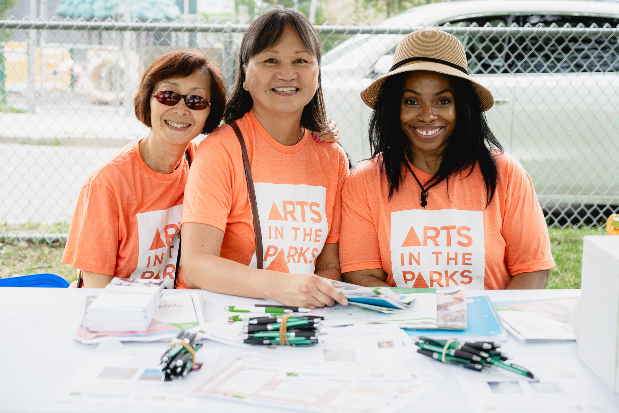 Three Arts in the Parks volunteers in orange t-shirts sit at the AITP booth. Photo by Kat Rizza.