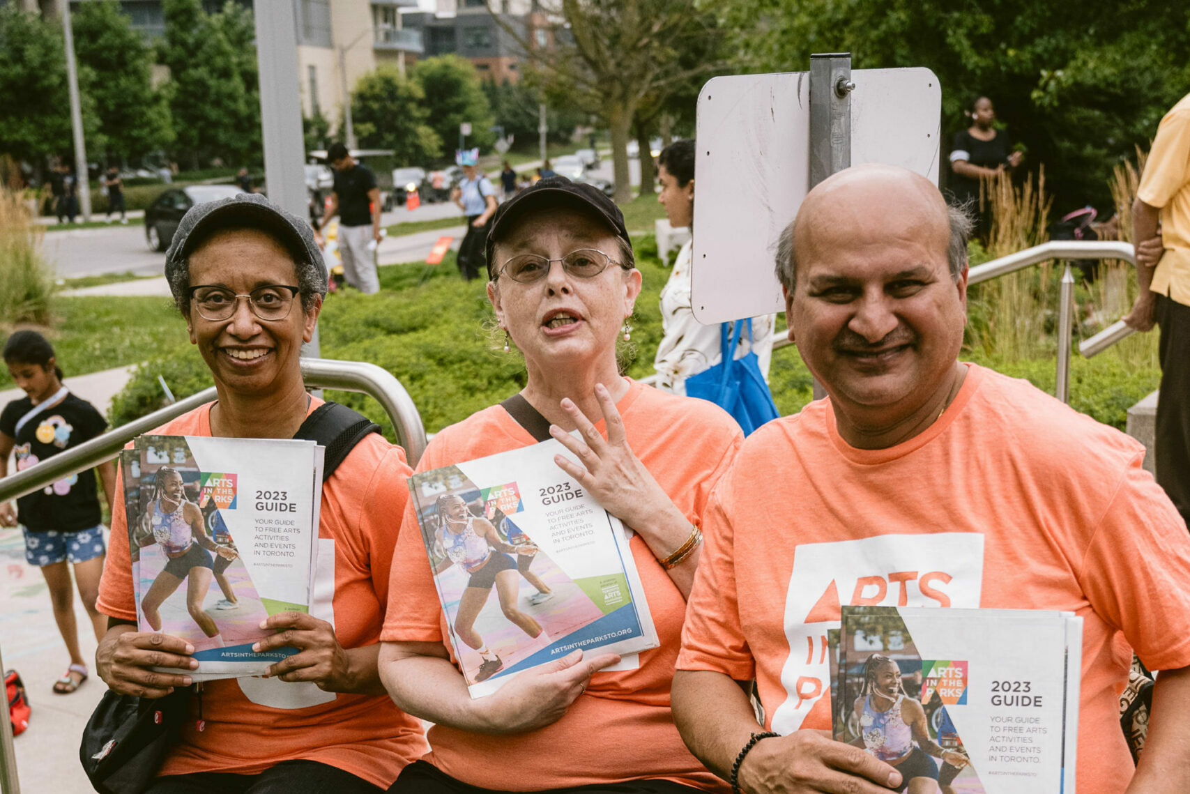 Three Arts in the Parks volunteers with orange t-shirts hold season brochures.