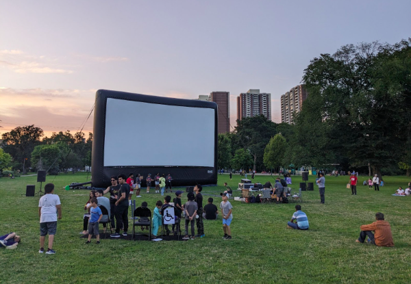 
A large inflatable screen at Dentonia Park. A crowd of people, mostly children, gather near the screen waiting for “English Vinglish” to begin. 
