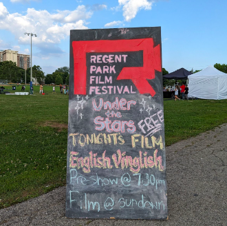 A chalkboard sign with Regent Park Film Festival’s logo in red, and the words “Under the Stars, Free. Tonight’s Film: English Vinglish. Pre-Show at 7:30PM, Film at Sundown”