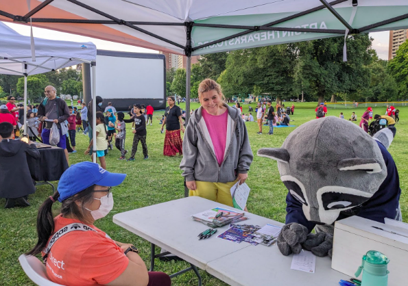 An Arts in the Parks Volunteer sits at a table while Feed Scarborough’s mascot, a raccoon, fills out an Arts in the Parks survey. 