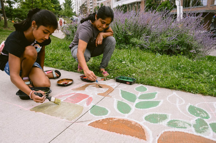 A women and a young girl crouch on the pavement and are painting a design of leaves using eco safe paint.