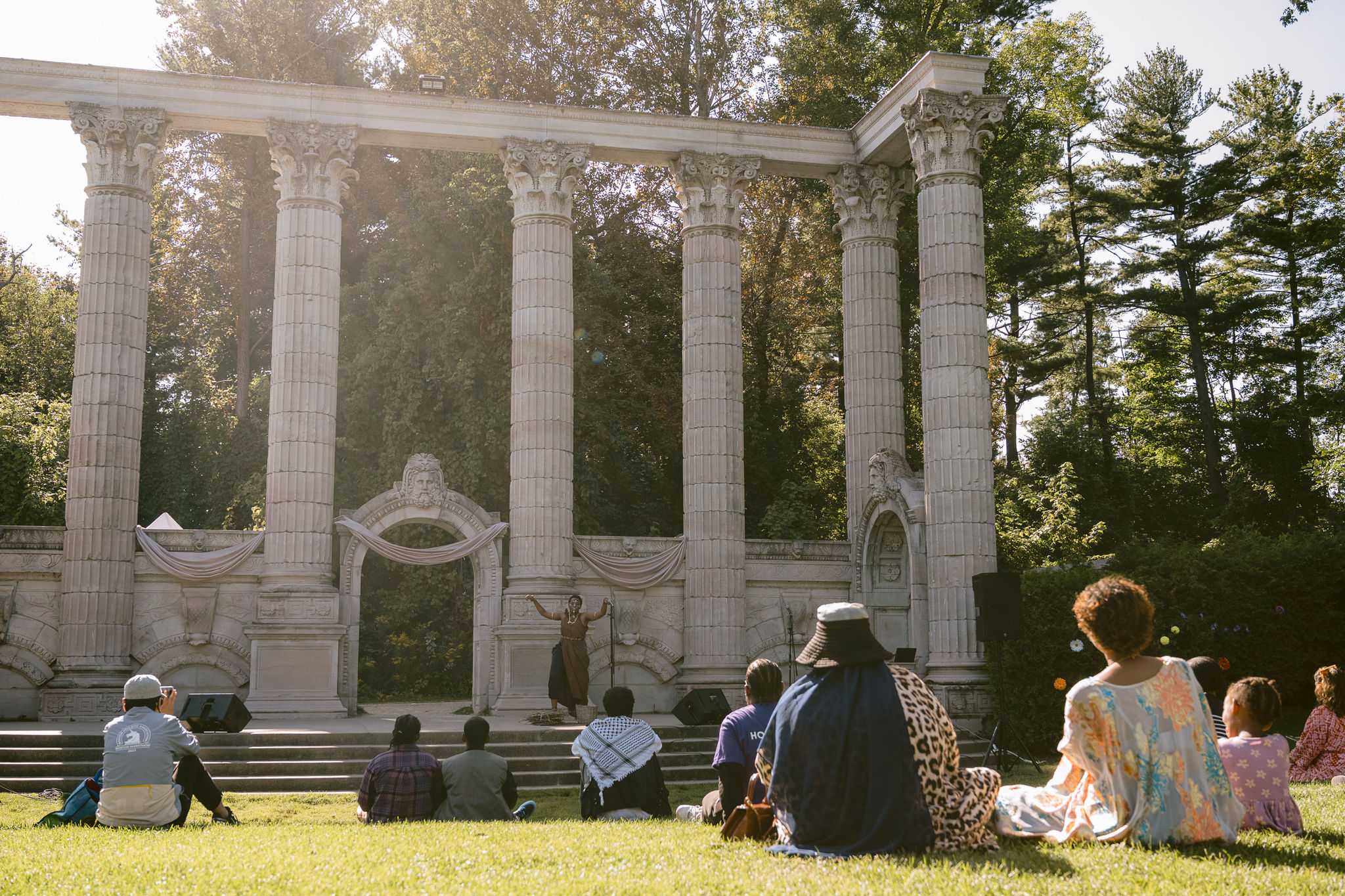 An audience sits in the grass to watch the performance on the outdoor stage surrounded by Greek-style columns