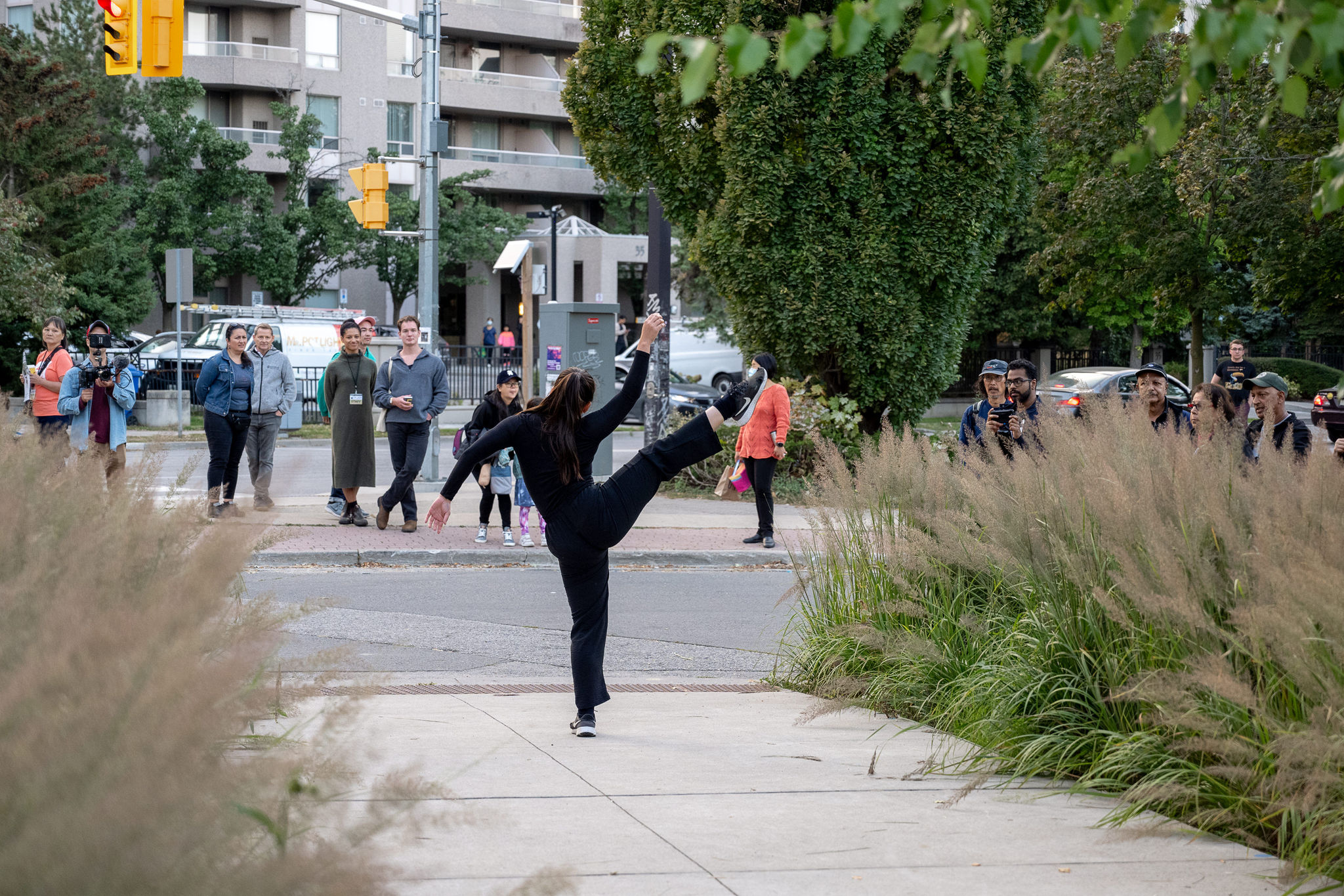 A woman dancer in black performs on a sidewalk as passersby watch.